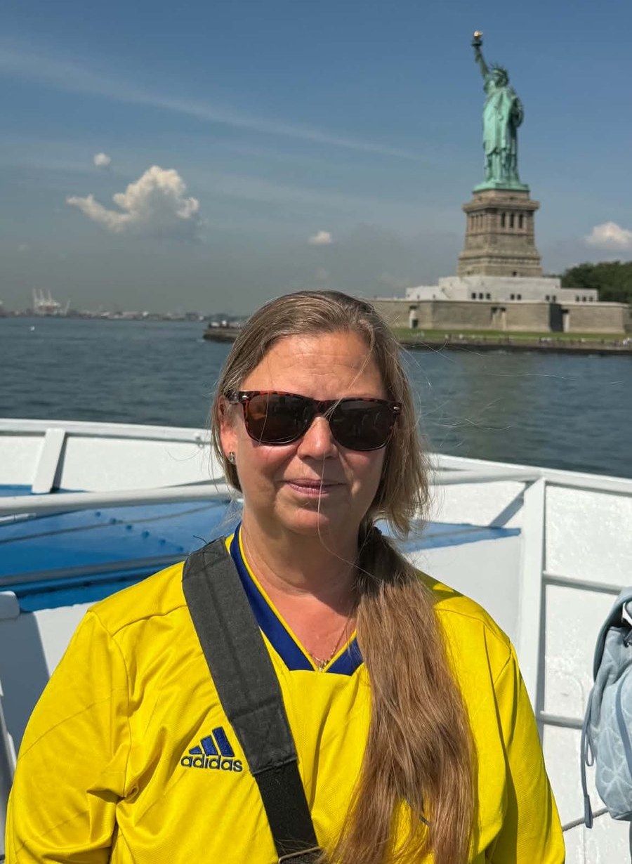 Susanne Petersson i gul landslagströja på en båt med Frihetsgudinnan i bakgrunden.